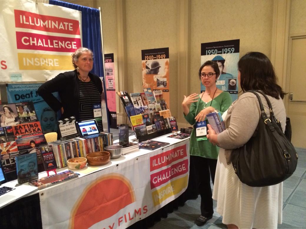 New Day filmmaker Regan Brashear stands behind the exhibition booth smiling as filmmaker Vanessa Warheit talks to a conference attendee.