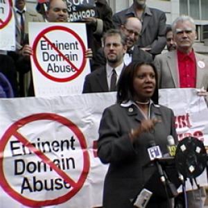 A Black woman in a suit stands in front of various news microphones making a speech. Behind her is a large banner that reads “Eminent Domain Abuse” with a large red slash through the words. A group of people of various races stands behind the banner, one holding a poster that has a red slash through the words “Eminent Domain Abuse.”