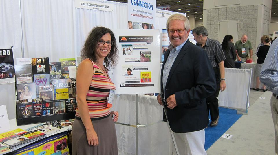 New Day Filmmaker Cindy Burstein and APA festival programmer Robert Simmermon smile for the camera by the New Day Films exhibition table. Conference attendees mingle around the booths behind him.