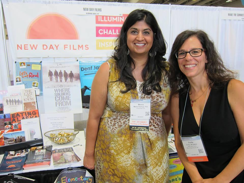 Meera Rastogi, APA member, and New Day filmmaker Cindy Burstein stand in front of the New Day Films booth at the APA conference and smile at the camera. They’re wearing professional clothing and conference badges are around their necks. The New Day Films booth includes a New Day banner, paraphernalia from various New Day films, and a bowl of candy.