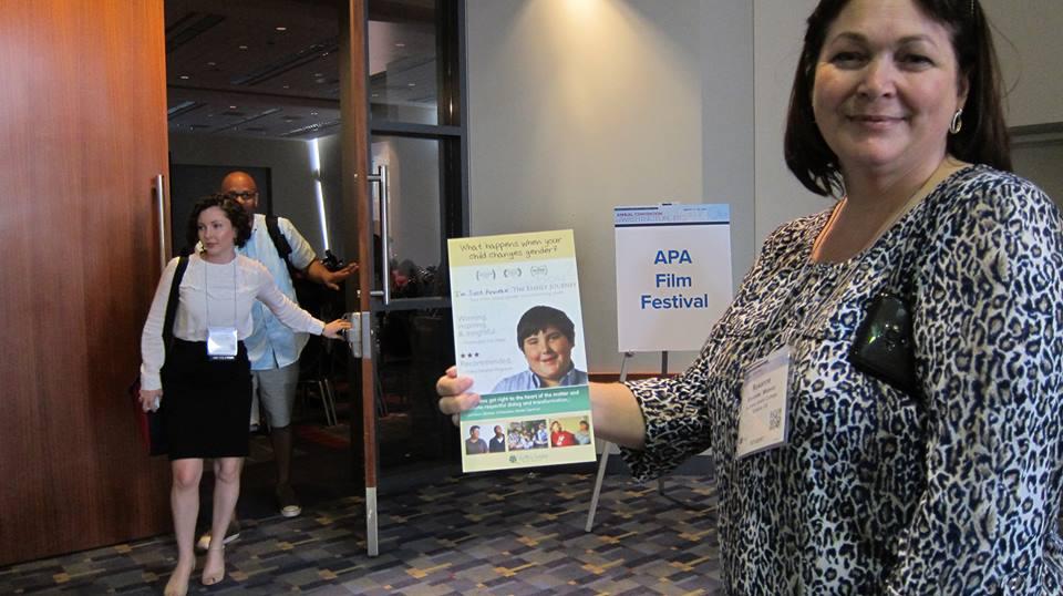A woman in a leopard print shirt with a conference badge around her neck smiles and holds out a flyer promoting the film “I’m Just Anneke.” Behind her stands a  poster reading “APA Film Festival” by the open door to a screening room.