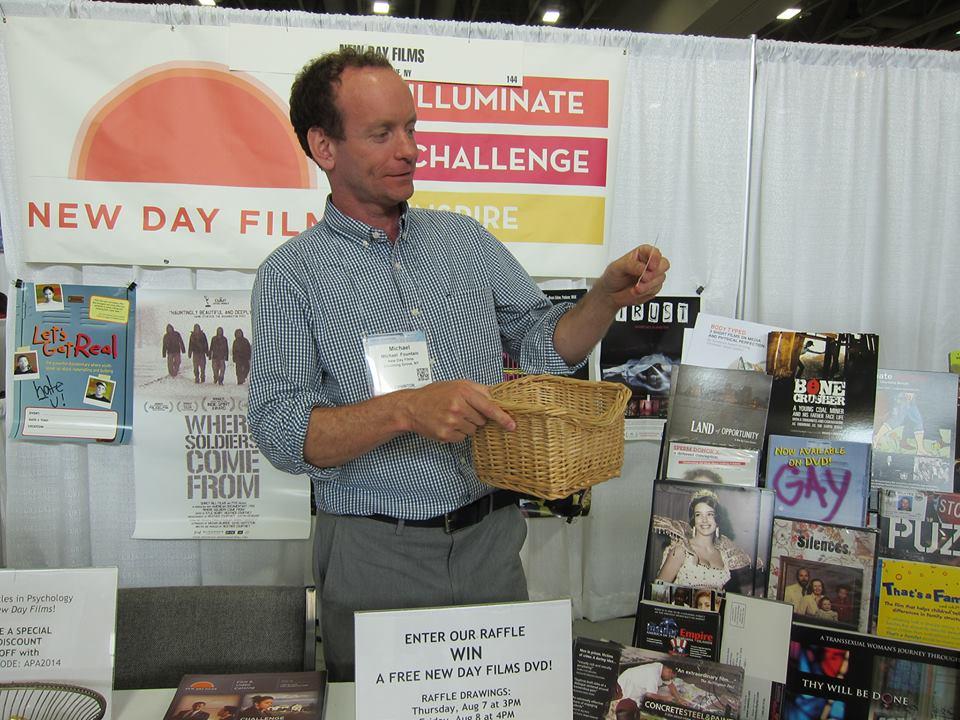 New Day filmmaker Mike Fountain wears his conference badge and stands at the New Day Films booth at the American Psychological Association conference with an amused expression on his face. He holds a wicker basket in his right hand and looks at a small slip of paper in his left hand.There is a sign on the table in front of him advertising a raffle. He is surrounded by promotional posters from various New Day films.