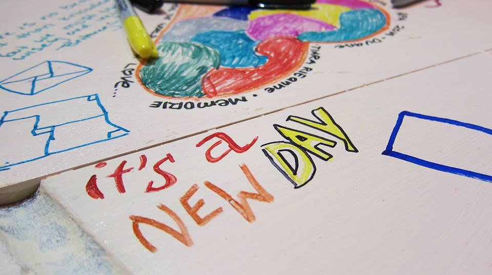 A close up of a white surface. The words “it’s a New Day” are written in sharpie in red, orange and yellow. Other shapes, designs, and writings cover other parts of the surface, including a big heart filled with different color patches and words around the outside such as “love” and “memories” On the edge of the heart rests a yellow sharpie with the cap pointing towards the words “it's a New Day”.