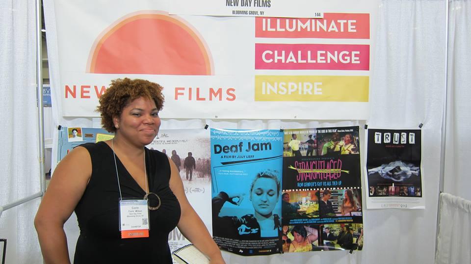 New Day filmmaker Corin Wilson wears a conference badge and smiles proudly at the New Day Films exhibition table. Behind Corin, a large banner for New Day Films with the logo and the words “Illuminate, Challenge, Inspire.” Below the banner are posters promoting various films from New Day.