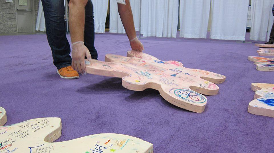 A person in black pants and orange sneakers wears clear plastic gloves and lays down an amoeba shaped piece of wood covered in sharpie writing and designs on a large purple carpet. On the edges of the image other similarly shaped pieces of wood covered in sharpie writings and designs are laid out on the purple carpet. Several white curtains hover slightly above the curtain in the background.