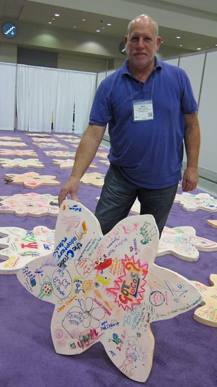 A white man in a purple t-shirt with a conference badge around his neck props up a knee-high piece of wood shaped like a flower and covered in colorful sharpie writing and designs. Rows of similarly shaped pieces of wood lay on the purple carpet in the background in rows. Rows of white curtains are hung up around the edges of the carpet to make a makeshift wall.
