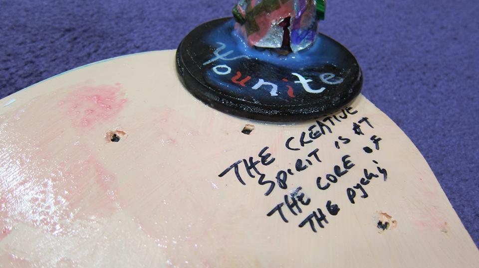 A close up image of a shiny painted wooden surface. On it, the words “The creative spirit is at the core of the psyche” are written in black sharpie. Above these words is a black circle with the word “Younite” written in red white and blue.