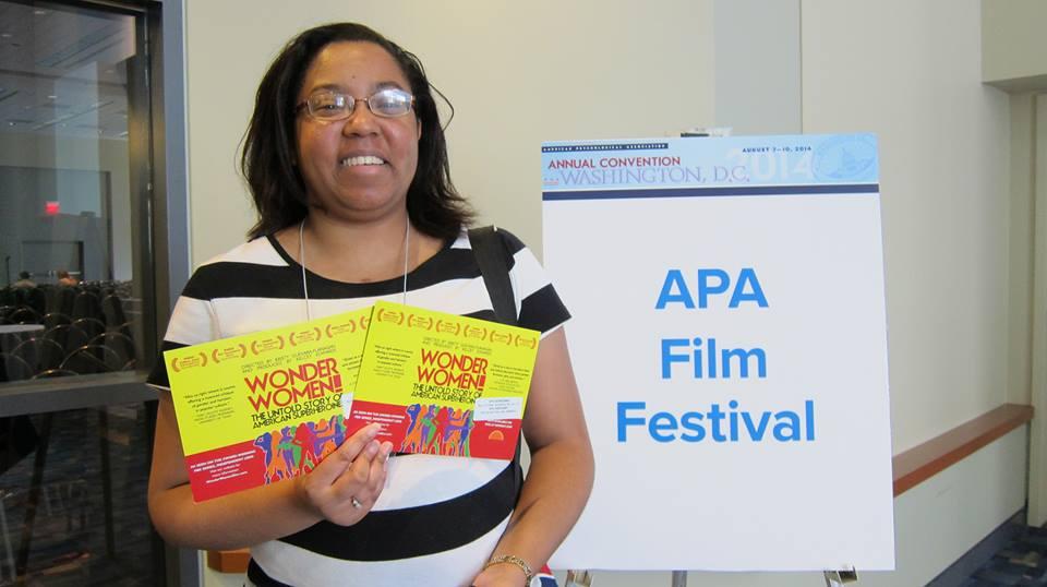 A woman holds two promotional flyers for the New Day film Wonder Women. Behind her is the APA Film Festival poster.