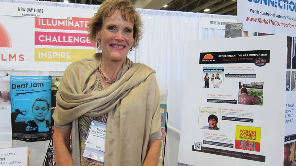 Mary Ann Dornbusch, APA convention manager smiles and stands in front of the New Day Films booth. To her right is a poster for the Screening at the APA Convention and behind her are different New Day promotional posters and banners.