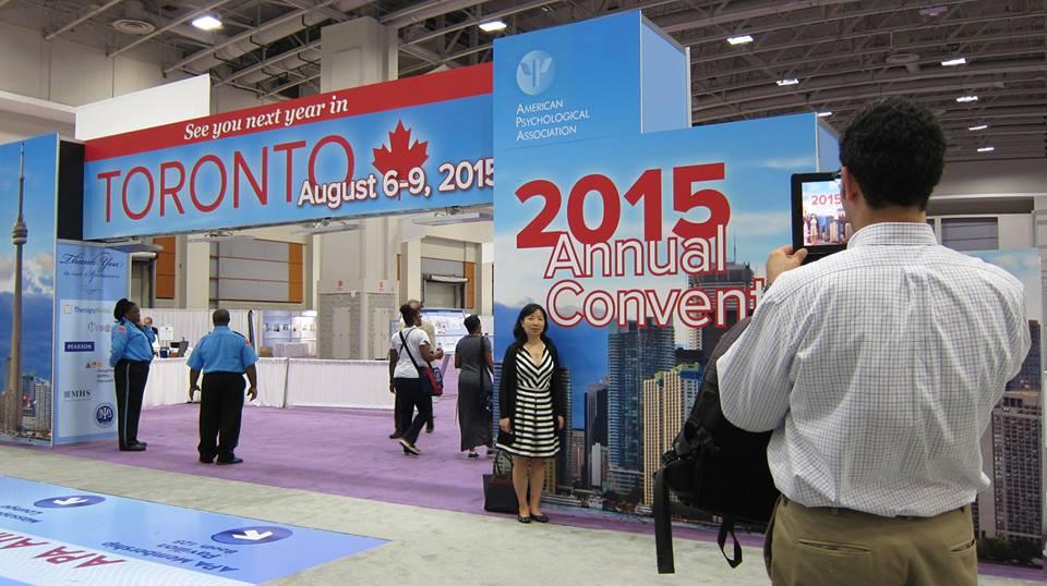 A woman smiles and stands in front of a large arch with the toronto cityscape printed on it. The top of the arch reads, “See you next year in Toronto August 6-9, 2015.” A man in the foreground takes a picture of her on an iPad.