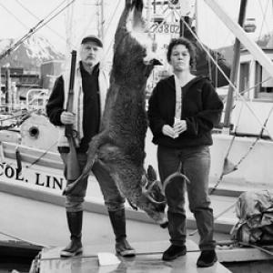A black and white still from the New Day film Eating Alaska. Two people with serious, somewhat angry expressions stand on a dock on either side of a dead animal hanging upside down. They are wearing long pants tucked into wellington boots and long sleeved tops. The person on the left wears a vest and holds a gun in his hand. The person on the right holds their hands together at their waist. Behind them is a commercial fishing boat.