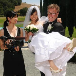 Asian-American New Day filmmaker Debbie Lum wears a black dress and holds a camera. She smiles and looks to the side at a young Asian woman in a bridal gown with a veil, a white bouquet and white high heeled shoes. An older white man is holding her and wearing a suit. The couple smiles at the camera. There is a garden behind the trio.