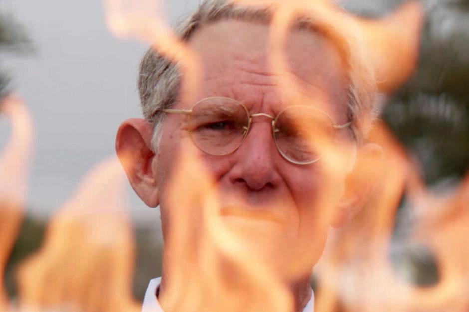 An older white man with glasses and a furrowed brow stands outside on a cloudy day. In front of him, orange-yellow flames rise up, partly blocking the view of him.