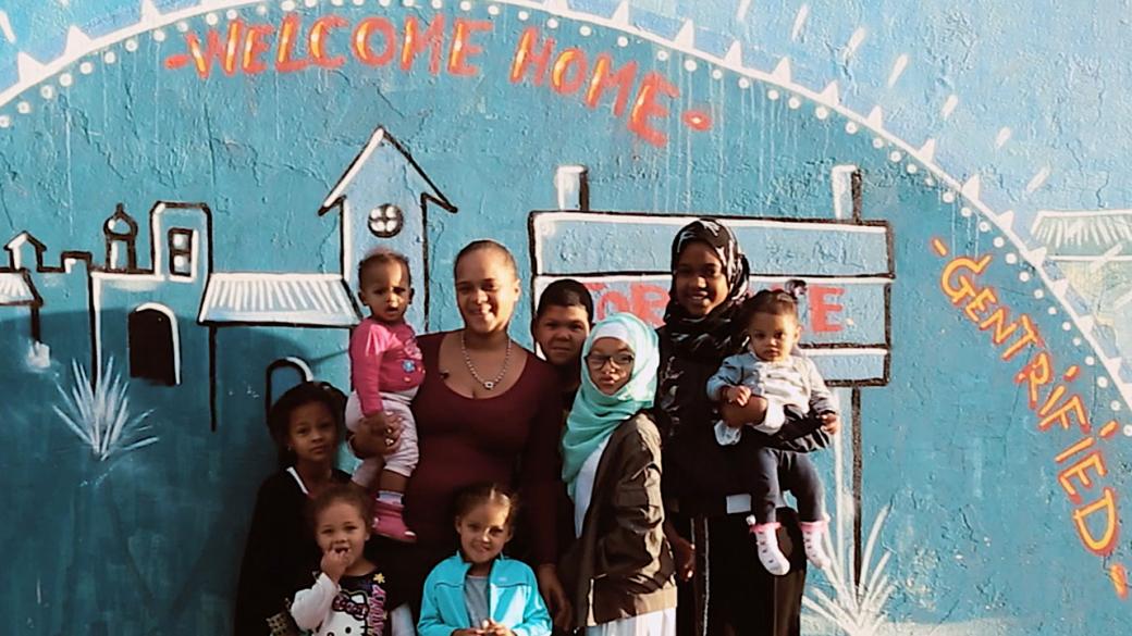 Two adults stand in front of a colorfully painted mural of buildings with "Welcome Home" and "Gentrified" painted above the buildings. They're smiling with six children.