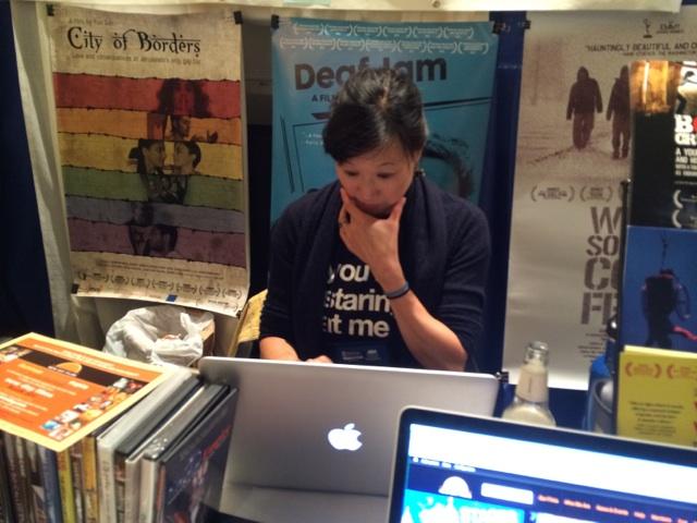 New Day filmmaker Debbie Lum focuses intently on her laptop at the New Day booth. She’s surrounded by promotional materials for various New Day films including posters for City of Borders and Deaf Jam.