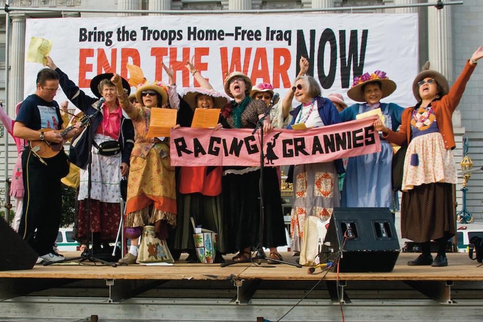 Seven older women wearing straw hats, and colorful aprons stand on a wooden stage and sing into microphones accompanied by a mandolin player. Several hold up a pink sign with black block letters reading, “Raging Grannies” and a silhouette of an older woman in a dress, brandishing an umbrella. Behind them, a giant banner reading, “Bring the Troops Home--Free Iraq. End the war NOW.”