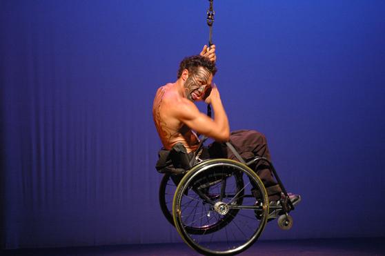 Rodney Bell, a Maori performer with facial tattoos is shirtless, sitting in a manual wheelchair attached to a rope by a harness. He floats in the air above a stage, a cobalt blue background behind.