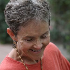 An image from the New Day film States of Grace. Dr. Grace Dammann closes her eyes and smiles towards the ground, her body tilted slightly. Her gray and white hair is short and she wears earrings, a necklace and a long sleeved shirt. The background is full of blurred out foliage.