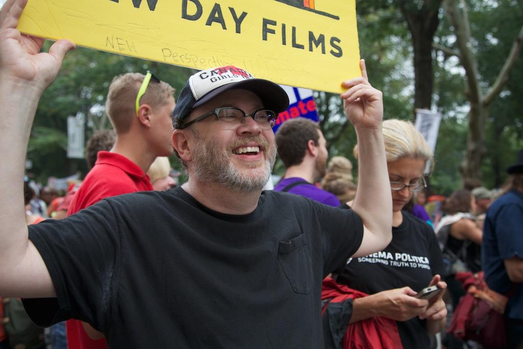 New Day filmmaker Jay Rosenstein looks off camera as he smiles and holds a yellow New Day Films poster on top of his head. He is wearing glasses, a black t-shirt and a baseball cap. A large group of people, some with climate protest posters mill in the background in front of a park with trees.