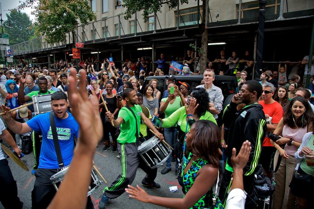 A large crowd of people on the street. Various young people of color wear bright green and blue clothing move forward in the street as they dance and play the drums, many with their mouths open in chant or song. One person’s raised hand is prominently visible in the foreground.