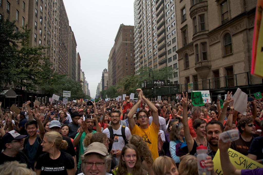 A massive group of people of various races, ethnicities and genders pack together in a city street. Many raise their hands towards the sky, clapping, making peace signs, taking pictures or holding signs up. The majority of people face forward as they move through the streets in a march. The tall city buildings rise up on both sides of the frame.