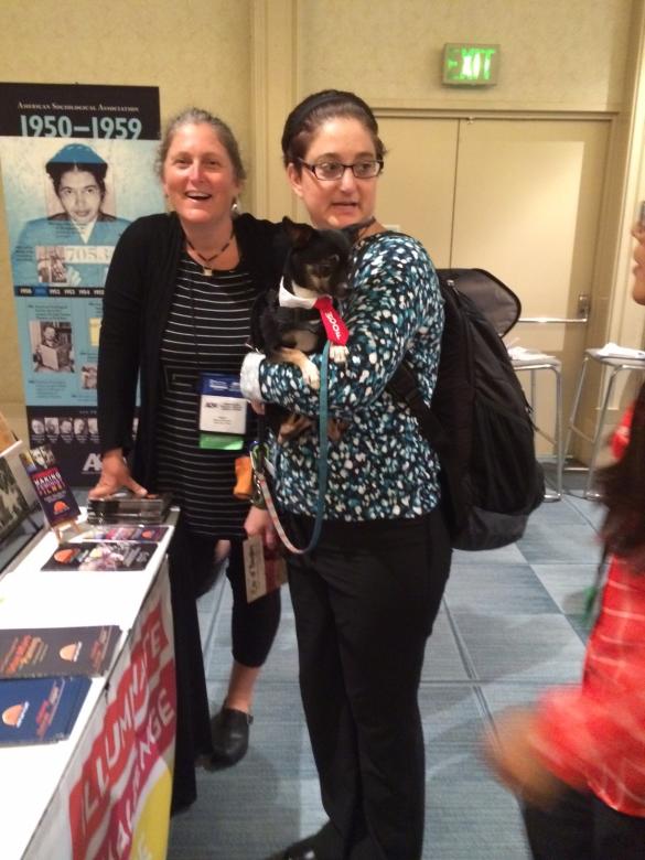 A conference attendee at the New Day Films booth holds a small service dog in a red tie with “woof” in white lettering.