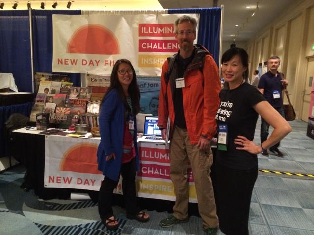 New Day filmmakers Yun Suh and Debbie Lum stand at the New Day Films exhibition table with a conference goer and smile for the camera. Debbie Lum wears a promotional t-shirt for her film seeking asian female, a black t-shirt that reads “you’re staring at me” in white text.