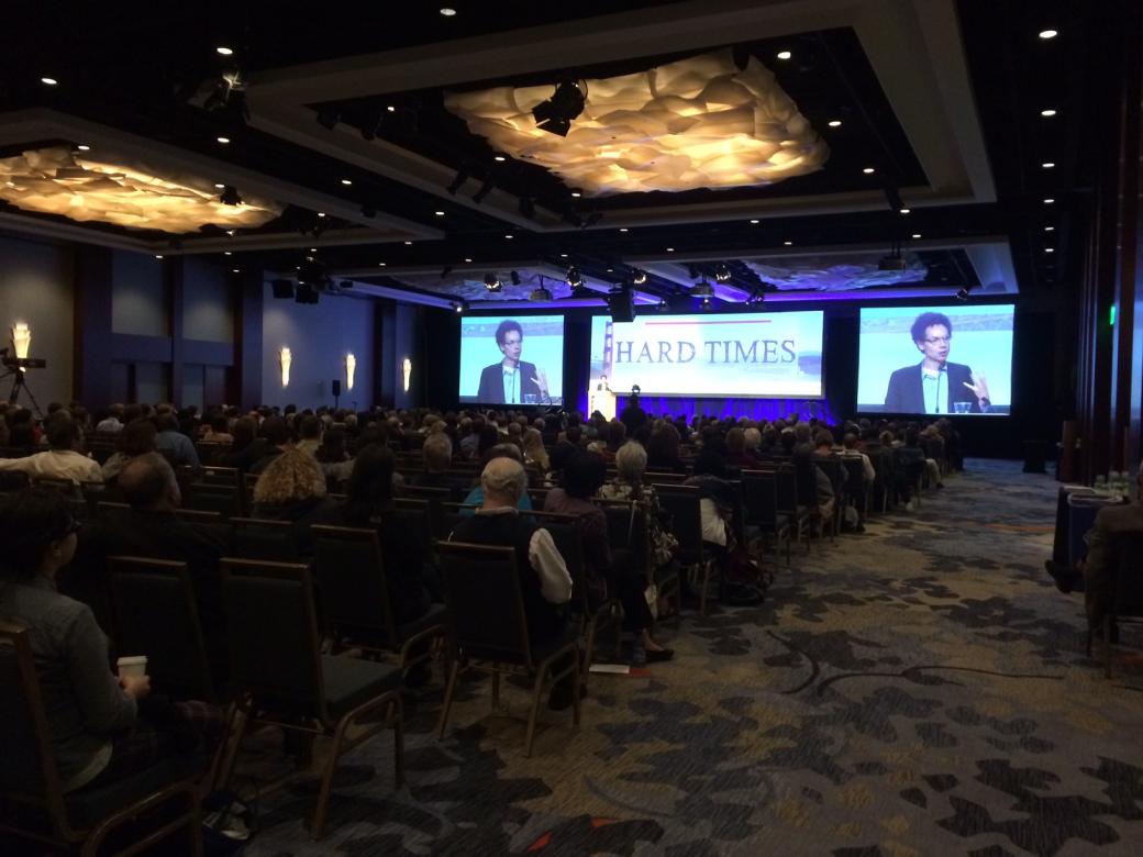 A large conference room with a mostly full audience watching keynote speaker Malcolm Gladwell at a podium in the front. Two large screens magnify his image as he speaks. Between the two screens is a larger screen with an image of the golden gate bridge and the words “Hard Times” in big letters.