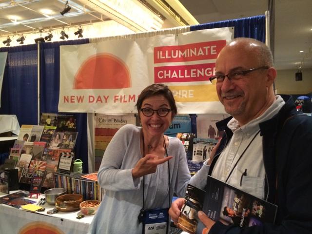 At the New Day Films exhibition booth, New Day filmmaker Vanessa Warheit smiles and points excitedly to Professor Ted Cohen and has one handon his shoulder. He holds a New Day catalog and smiles.