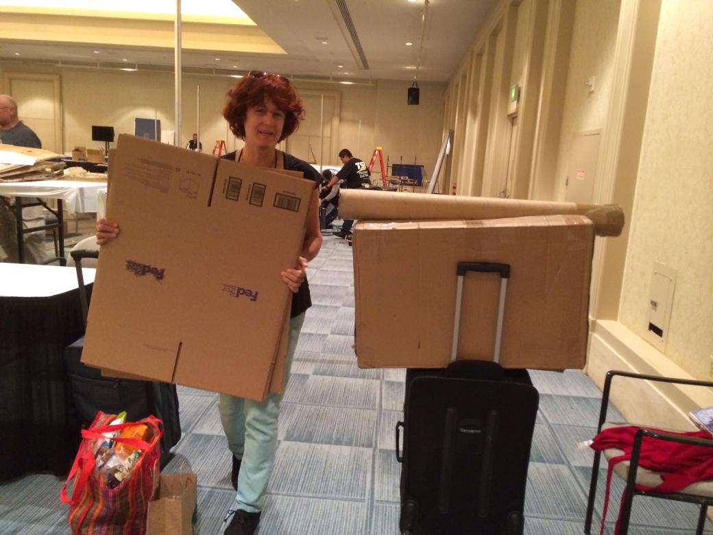 New Day filmmaker Frances Nkara packing up the New Day Films booth into cardboard boxes and suitcases. In the background are a few ladders, cardboard bags, empty conference tables, and other people break down their booths.
