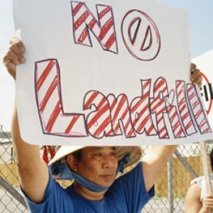  A still from the New Day film A Village Called Versailles. A Vietnamese person holds up a sign that reads “No Landfill” in large red and white striped letters. The “O” in “no” is a circle with a slash through it. They are wearing a conical hat tied under their chin with a large blue ribbon.