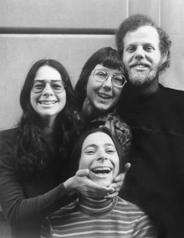 A black-and-white photo of New Day Films’ founders. Amalie Rothschild, Julia Reichert, and Jim Klein stand behind a seated Liane Brandon. Amalie cradles Lian’s face, and they all smile playfully.