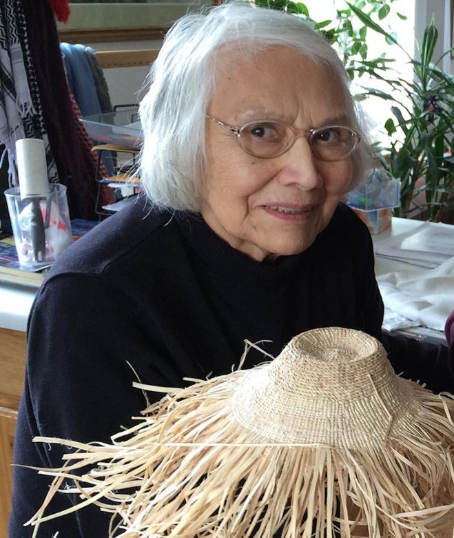 Haida elder and weaver Delores Churchill smiles at the camera. She is wearing glasses and a black top. She holds up a weaving work in progress with the unfinished ends splaying out.