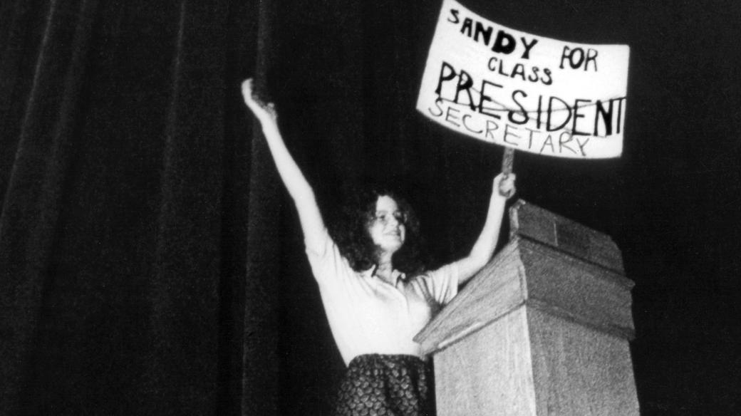 In this black and white still from the film, a young woman triumphantly holds up a sign that reads “Sandy For Class President.” The word ‘President’ is crossed out and underneath is written “Secretary”. She’s onstage at a lectern.