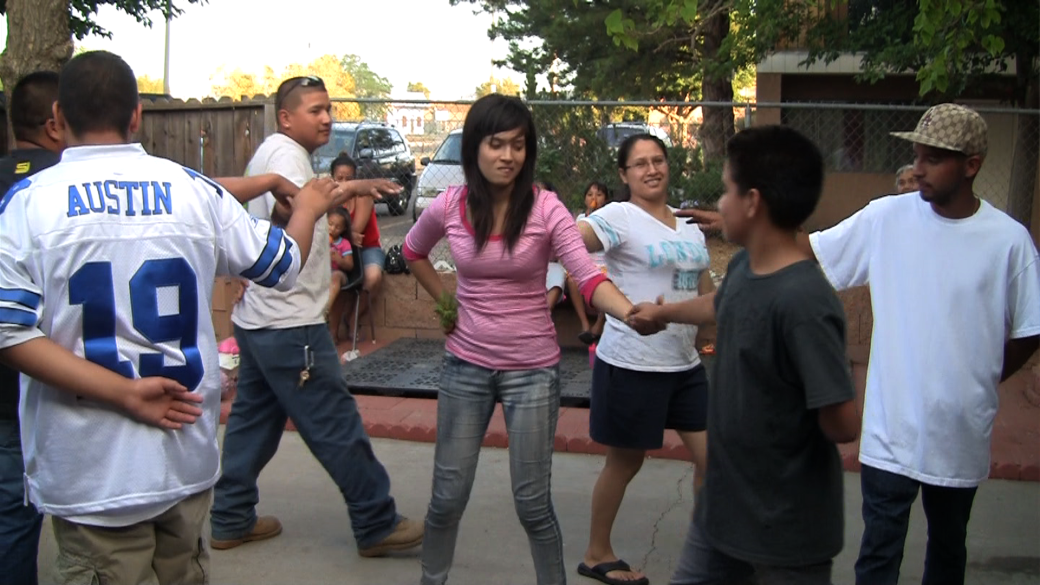 A group of seven people, mostly young, are dancing. They hold one arm outstretched and the other behind their backs. In the center, a girl and a boy are holding hands.