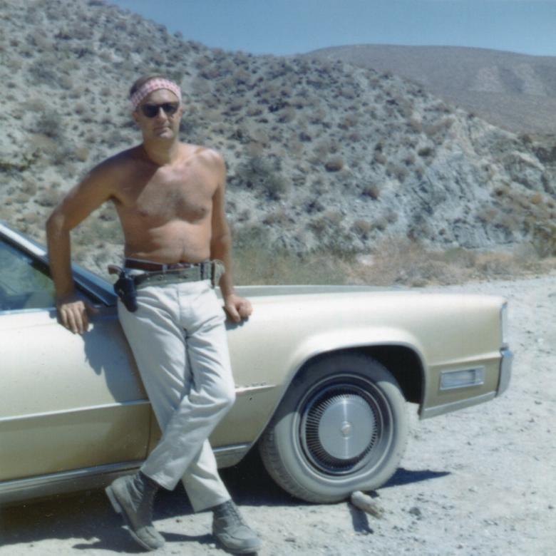 A still from the New Day film The Self-Made Man. An old color photo with off-white edges like a polaroid. Bob Stern, a shirtless man in well worn jeans and sunglasses leans against the side of a tan car from the 70s. The car is on a dusty, dirt road and there are mountains in the background filling the rest of the frame.