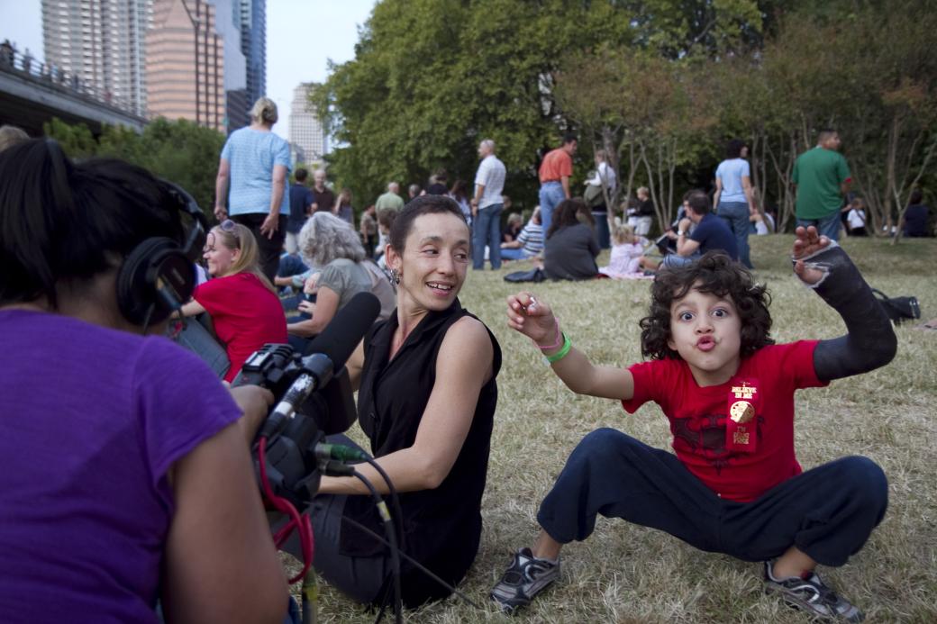 People sitting in the grass at a park. A child squats like a frog, arms raised, and making a goofy face. One arm is in a cast. A woman looks at the child, smiling. Another person is filming the two of them.
