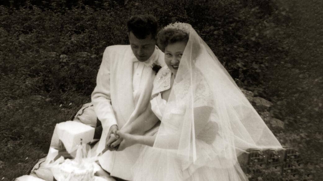 In this aged black and white photo, a bride and groom wearing white are sitting outdoors while jointly cutting a white wedding cake.