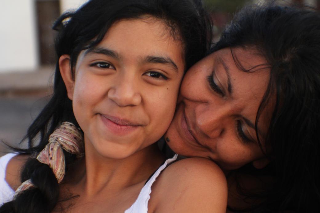 A still from the New Day film Life on the Line. A young Mexican-American girl, Kimberly Torrez smiles sweetly towards the camera. A woman, likely her mother Vanessa Torrez sits behind Kimberly and rests her head on her shoulder. She turns her face into Kimberly’s neck with a loving smile.