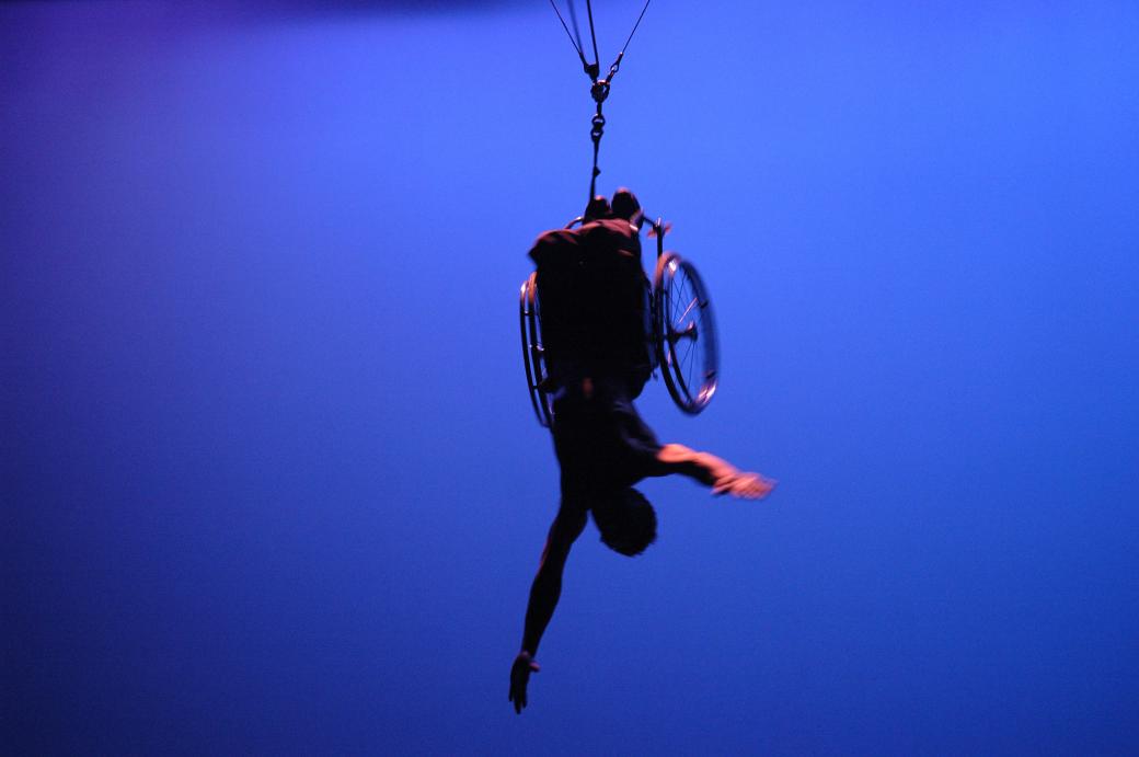 A person in a wheelchair suspended upside down with their arms extended, against a bright blue background.