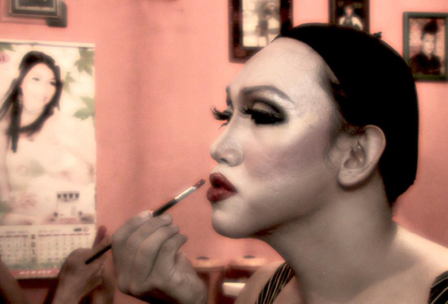 Profile image of a transgender woman in Indonesia. She focuses on her face in the mirror as she paints her lips. She has dramatic eye makeup and her hair pulled back under a cap.