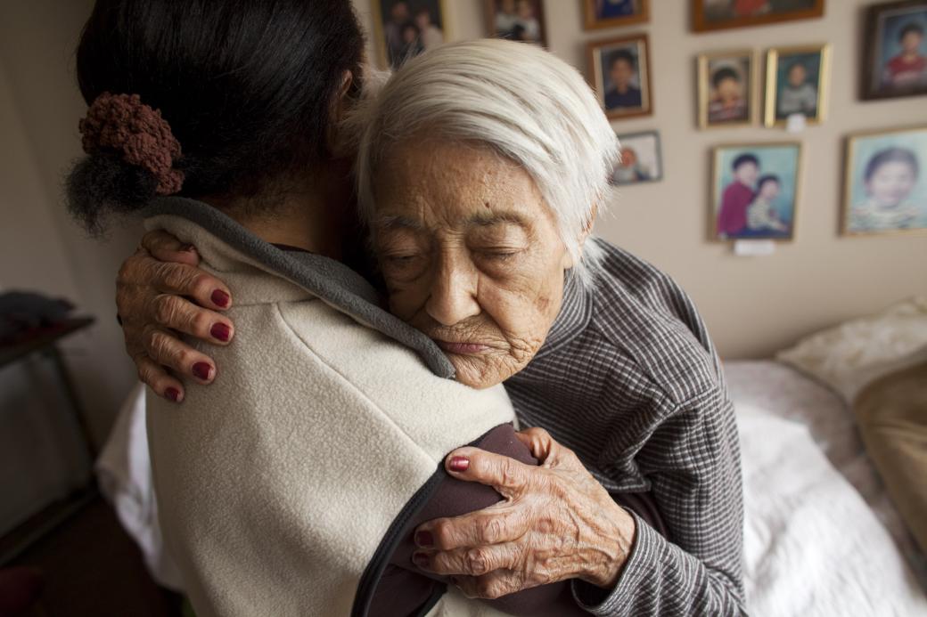 A still from the New Day film The Caretaker + The Mayor. An elderly woman with white hair hugs a person who is facing away from the camera. Her eyes are closed and her expression is vulnerable as she holds the person close. Behind them is a wall full of framed photos of a young child.