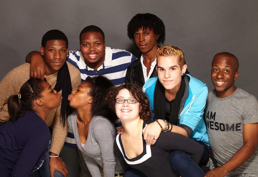 A group of queer teenagers having fun together. They are mostly Black youth. Two girls pucker up to kiss. The boys behind them have their arms around each other. One kid rides on another girl’s back. One kid wears a t-shirt that says I Am Awesome.