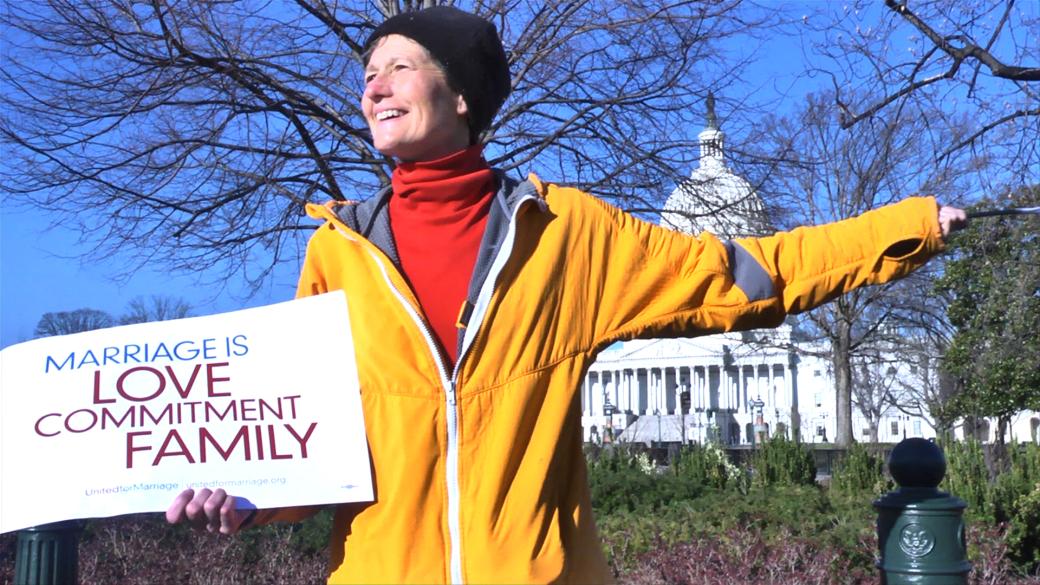 A white woman stands outside in front of a bare tree with blue sky. She smiles broadly, looking to the side. She is holding a white sign that reads “Marriage is Love, Commitment, Family” in blue and purple text. She wears a bright yellow zip up jacket which is half-unzipped to reveal a bright red turtleneck underneath and a black beanie on her head.