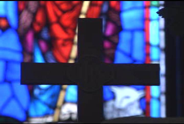 In front, the silhouette of a cross. Behind and out of focus, a blue, white, purple and red stained glass window.