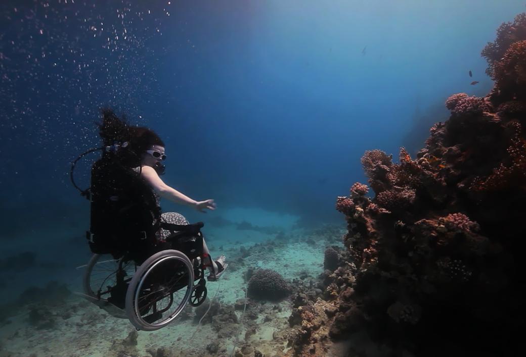 A still from the New Day film Fixed: The Science of Human Enhancement. A woman in a wheelchair is scuba diving. She floats slightly above the ocean floor. Her arms are bare and she does not appear to be wearing full scuba gear other than small goggles. Her hair drifts up in the water and she holds her arms out to the side for balance.