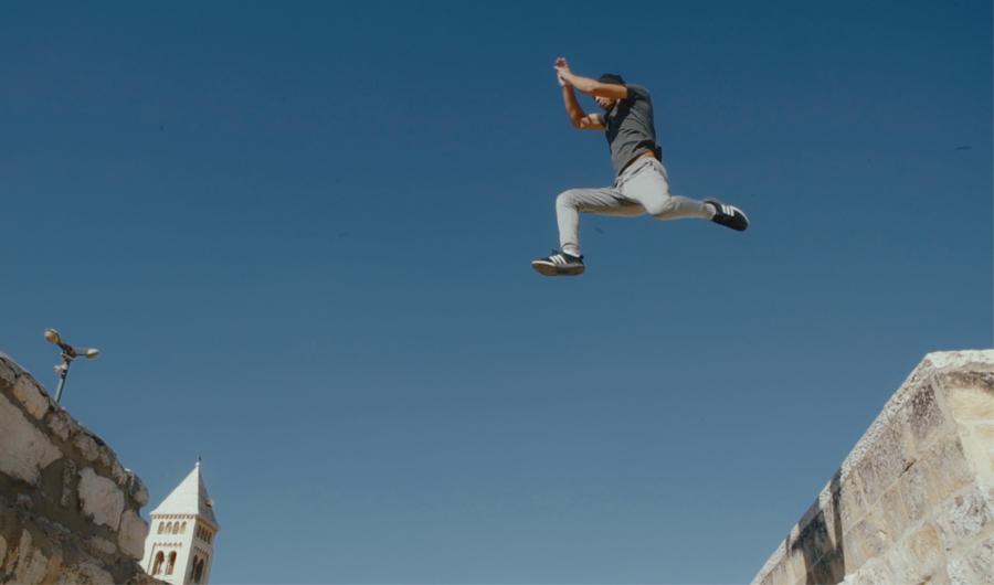 In this low angle shot, the camera is pointed up at a young Palestinian man leaping through the air as he jumps from one building to another.