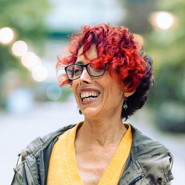 a short bright red and brown haired person wearing black square glasses and a V neck yellow shirt stands against a blurry background laughing