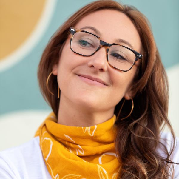 An auburn-long haired woman smiles, looking off-screen with her head tilted. She's wearing a yellow bandana and white t-shirt.