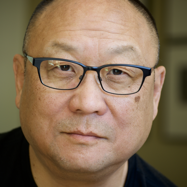 A medium shot of a man fills up most of the frame, with a blurred out background. He wears a blue shirt and thick rimmed glases.